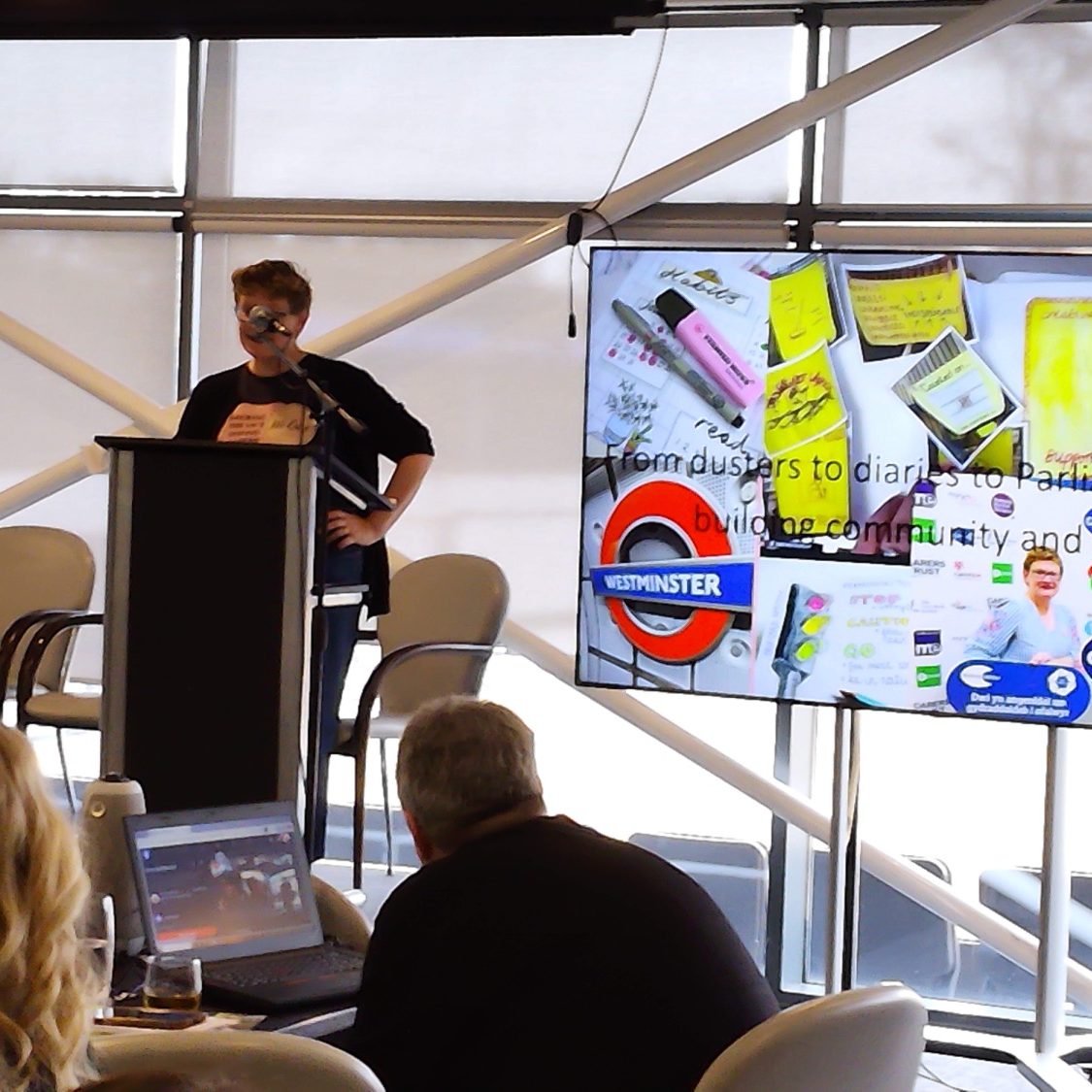 A speaker presents at a conference, with a screen displaying various documents and graphics.