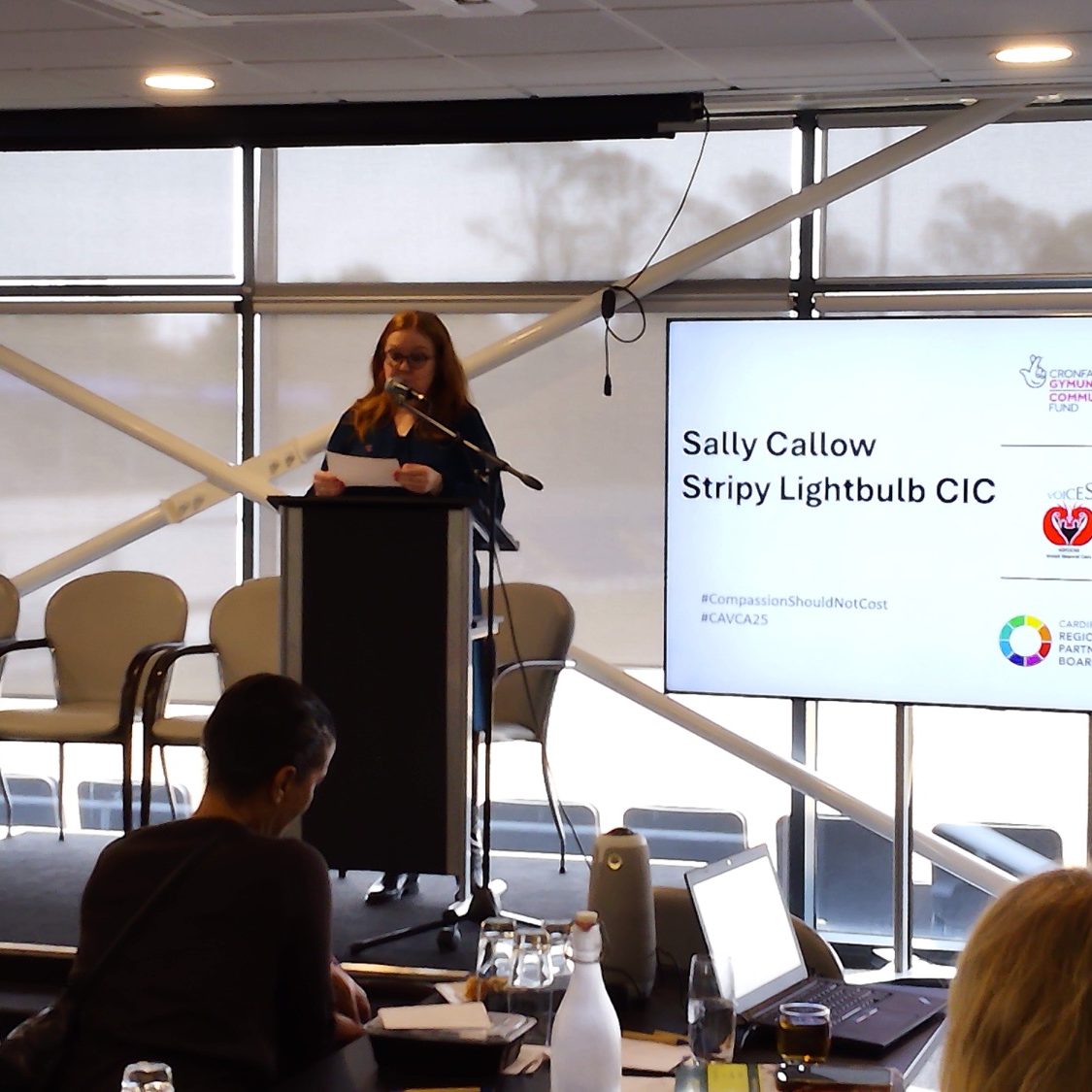 Woman speaking at a podium during a conference, slides visible in background.